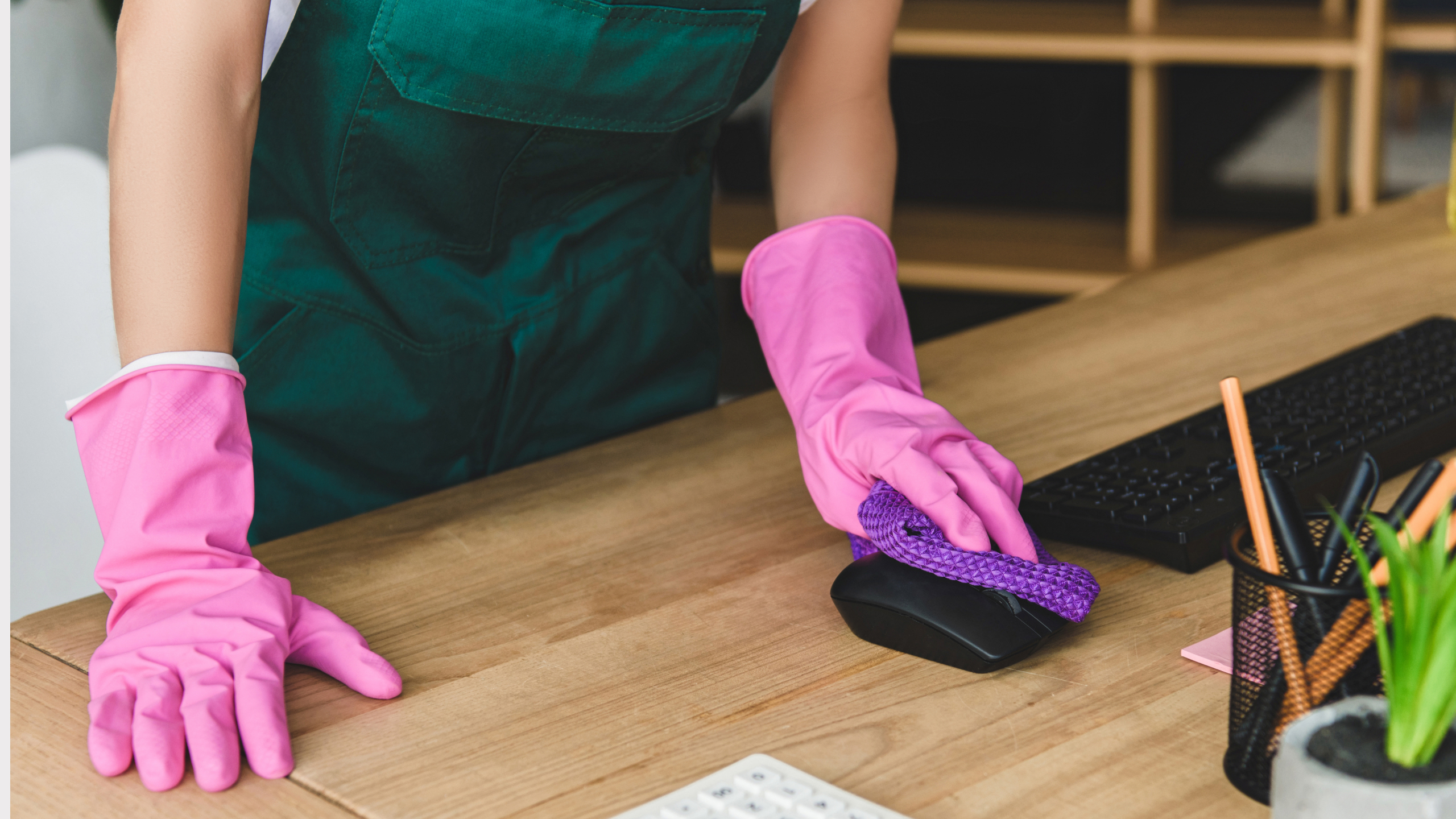 A person wearing pink rubber gloves is cleaning a computer mouse on a wooden desk with a purple cloth.